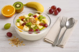 Glasschale mit Obstsalat und Quark, garniert mit Mandeln und frischen Früchten auf weißem Holztisch