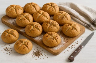 Frisch gebackene Kümmelbrötchen auf weißem Holztisch, bestäubt mit Mehl und Kümmel, daneben Tuch und Messer
