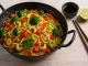 Buntes Wok-Gemüse mit Brokkoli, Paprika, Lauch, Sojasprossen und Nudeln, serviert im schwarzen Wok mit Cashewkernen auf hellem Holztisch