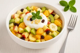 Obstsalat mit Äpfeln, Birne, Banane, Kiwi, Mandarinen und Skyr in einer weißen Schüssel auf hellem Holztisch