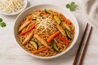 Asiatisch gebratene Nudeln mit Hähnchenstreifen, Paprika, Zucchini, Möhren und Sojasprossen in einer weißen Schale auf Holzhintergrund