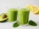 Grüner Smoothie mit Spinat, Avocado und Banane in Gläsern auf weißem Holzhintergrund serviert