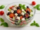 Glasschüssel mit mediterranem Käsesalat aus Feta, Tomaten, Gurken, Oliven und Kräutern auf weißem Holztisch mit Maserung